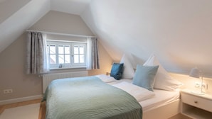 Une chambre à coucher avec un lit, des oreillers, une table de chevet, une lampe, une fenêtre avec des rideaux et un radiateur.