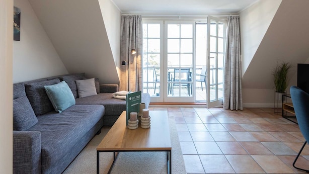 Un salon moderne avec un canapé gris, une table basse en bois et une vue sur l’extérieur grâce à de grandes fenêtres.