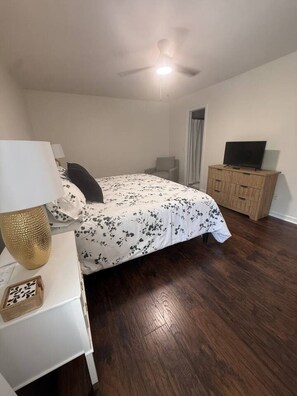 Ein Schlafzimmer mit einem Bett, einem Fernseher auf einer Holzkommode, einem Deckenventilator und einem Nachttisch mit Lampe.