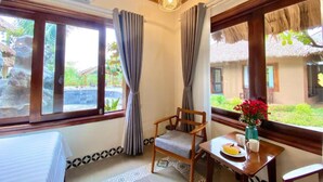A room with a bed, a chair, a table with a vase of flowers, and a view of a thatched-roof building. - Lemon Tree Homestay Tam Coc (Nam Hoa Lu)