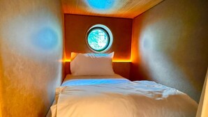 A narrow, dimly lit room with a single bed, a circular window, and warm lighting. - Stingy Hostel (Bangkok)