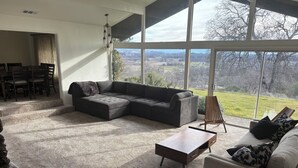 Un salón amplio con un sofá modular, una mesa de centro y vistas a un paisaje verde a través de grandes ventanales.