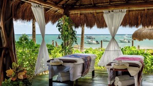 Dormitorio exterior con techo de paja, dos camas y vista al océano, con barcos al fondo.