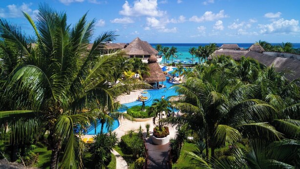 Un resort con piscina central, palmeras y edificios con techo de paja.