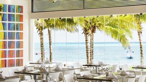 Un restaurante con mesas y sillas, ventanas amplias con vista al mar y palmeras, y coloridas obras de arte en la pared.