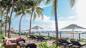 Un área de descanso frente a la playa con sillones y sombrillas, palmeras y vista al océano.
