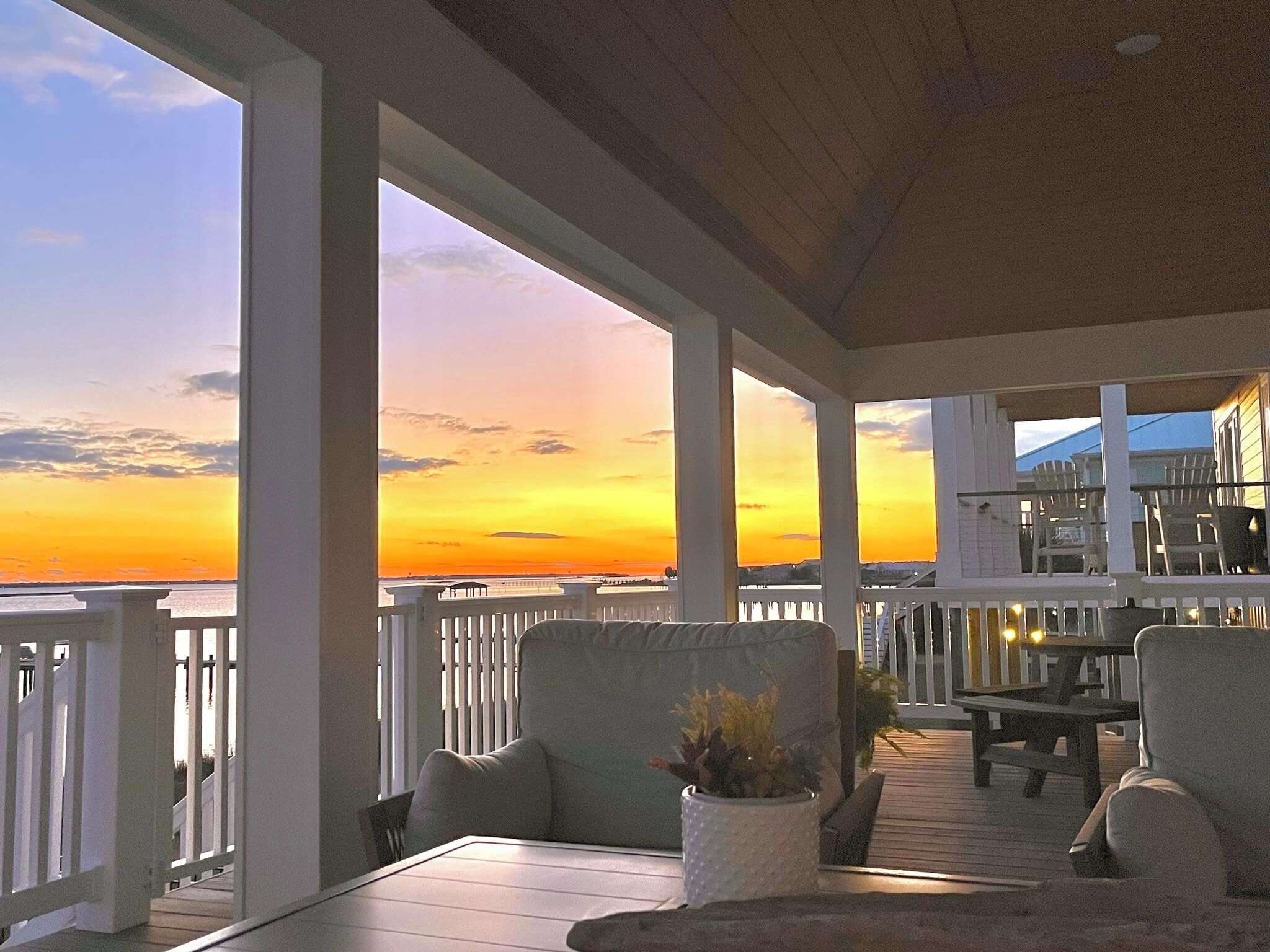 A covered outdoor area with a view of the sunset over the water, featuring a table and chairs, and a potted plant.