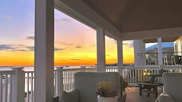 A covered outdoor area with a view of the sunset over the water, featuring a table and chairs, and a potted plant.