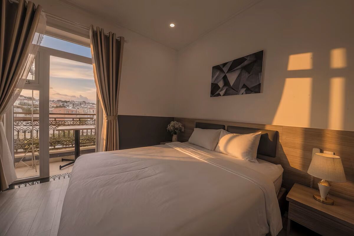 A hotel room with a bed, a nightstand, a lamp, a painting, and a view of a balcony with a railing.
