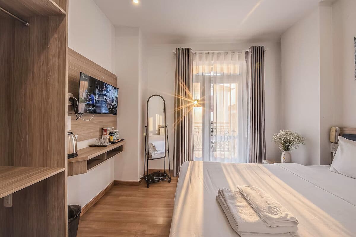 A hotel room with a bed, a TV, a mirror, a desk with a kettle, and a window with curtains.