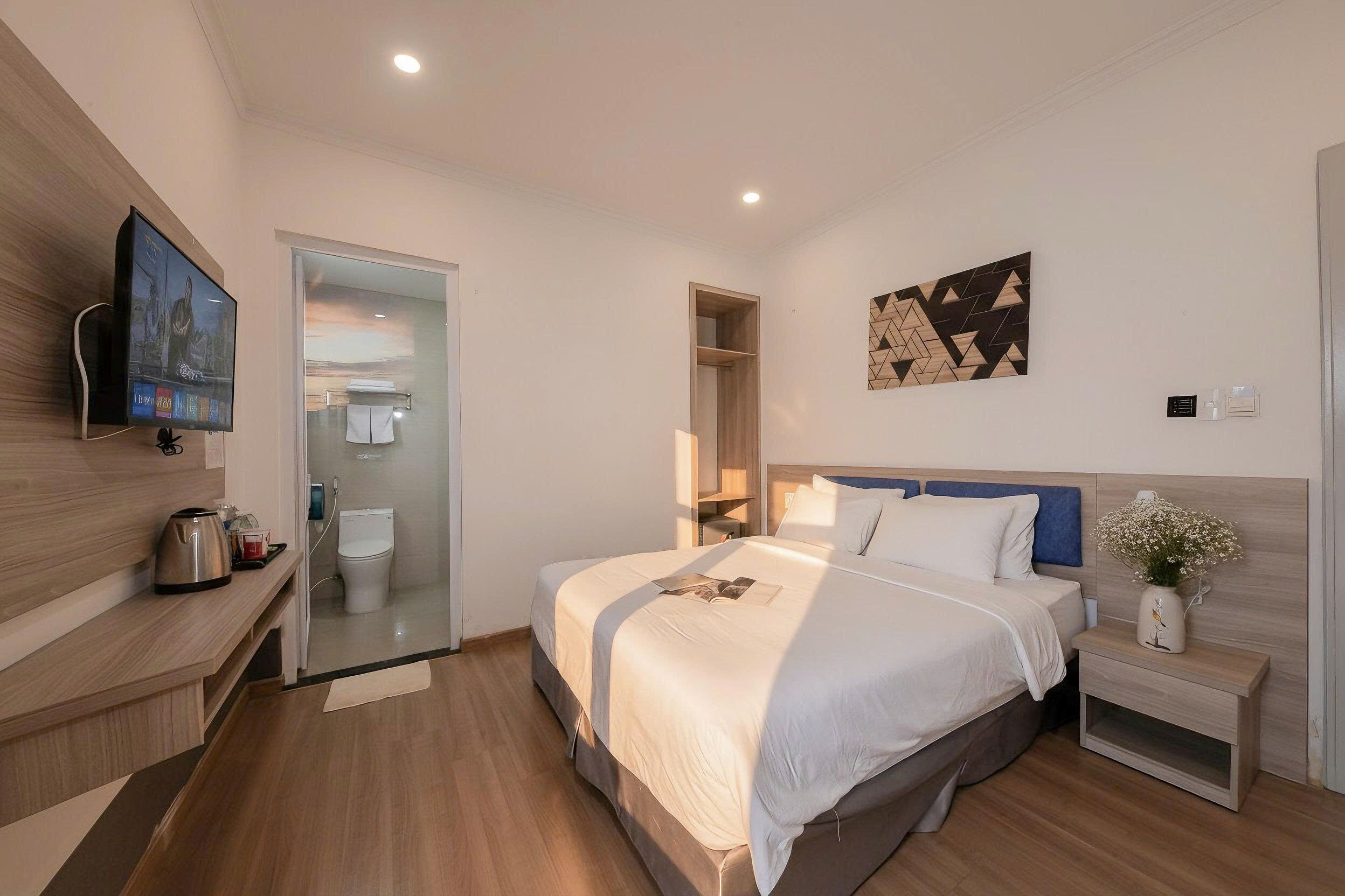A modern hotel room with a bed, bedside table, TV, and a bathroom visible through a glass door.