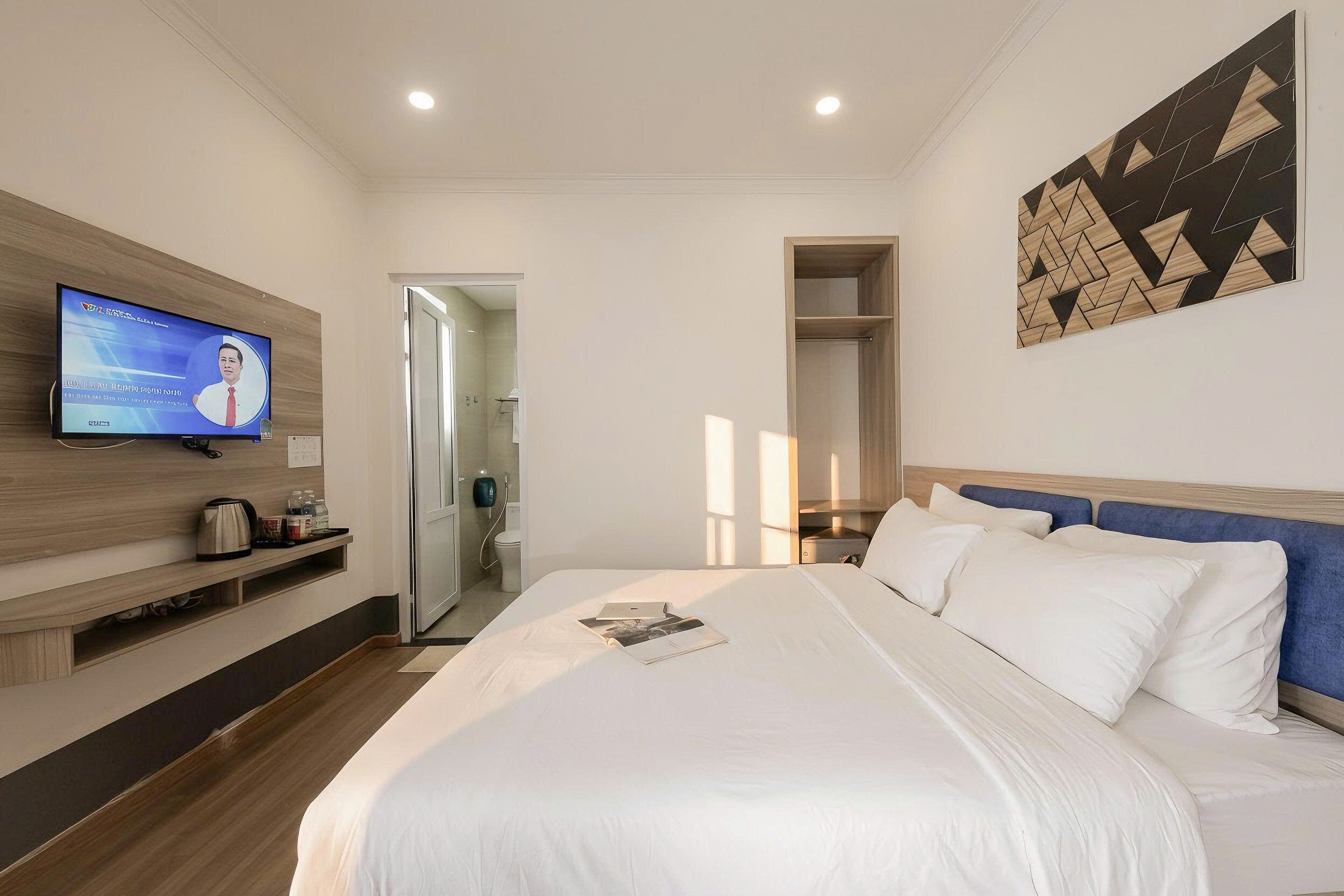 Quarto de hotel com uma cama, uma televisão na parede, uma prateleira de madeira e uma porta para um banheiro.