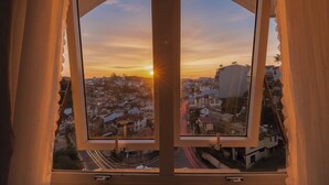 Vista a través de una ventana con marco de madera, mostrando un atardecer sobre una zona residencial con casas y una calle.