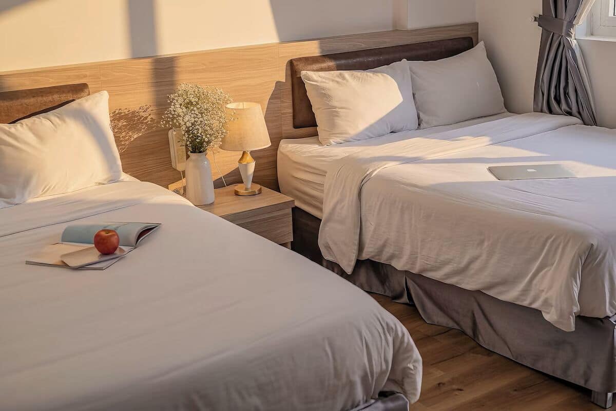 A hotel room with two beds, a nightstand with a lamp, a vase with flowers, and a laptop on one bed.