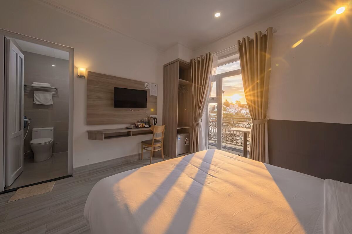 Habitació d'hotel amb llit, televisió penjada a la paret, escriptori amb cadira, bany i un balcó amb cortines.