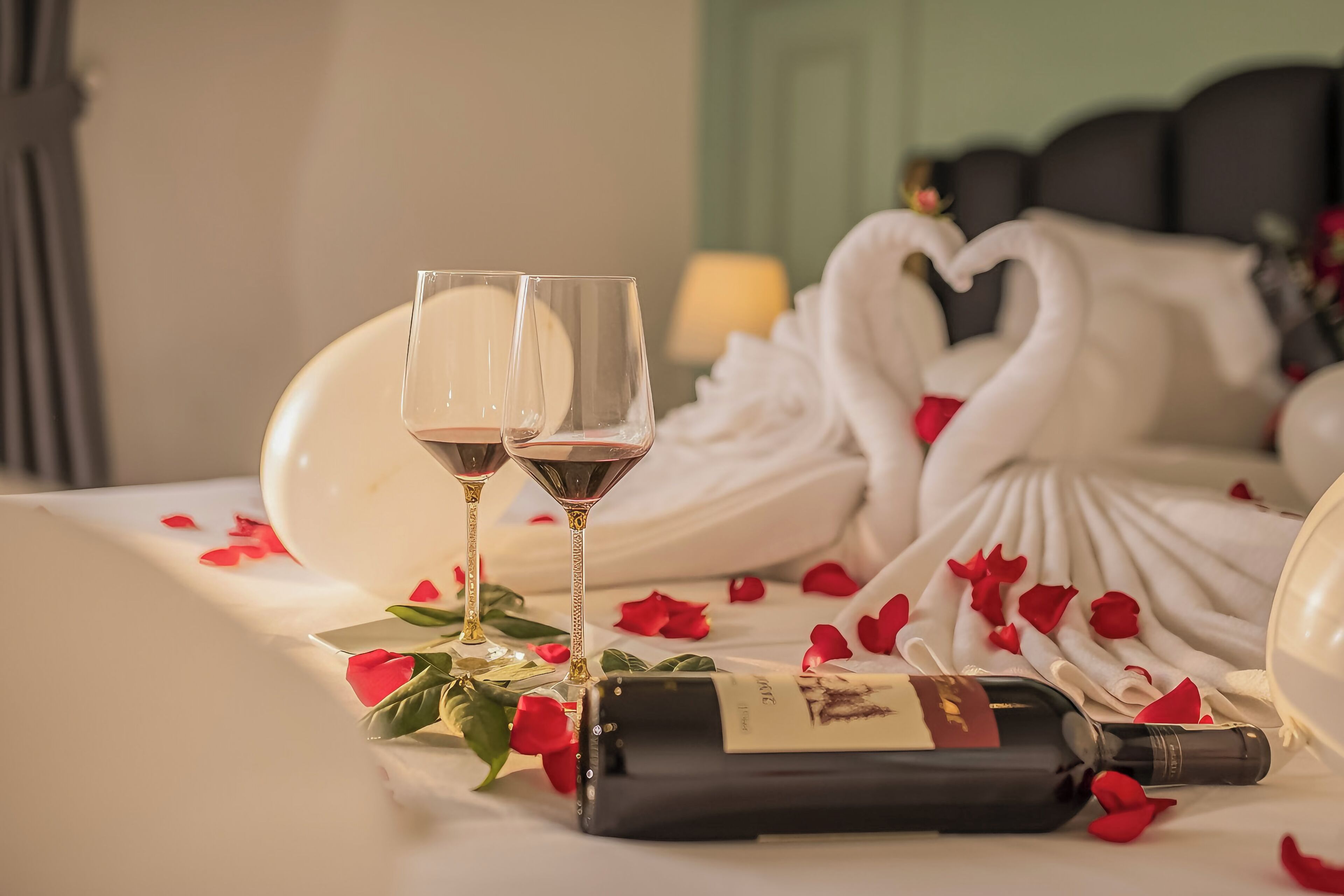 A bed with rose petals, two wine glasses, and a bottle of wine.