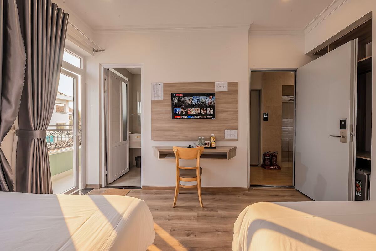 A hotel room with two beds, a desk with a chair, a TV mounted on the wall, and a view of the outside balcony.