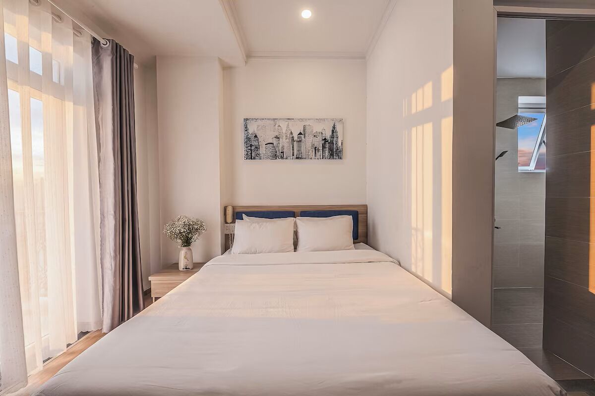 A hotel room with a bed, a nightstand, a vase with flowers, and a cityscape painting on the wall.