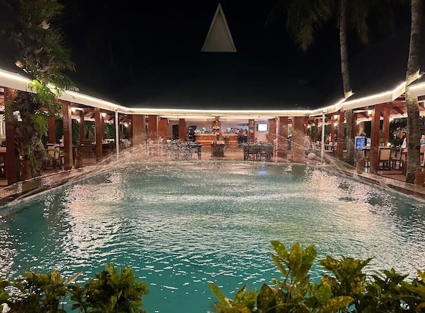 A nighttime view of a hotel pool area with a covered seating area and a bar.
