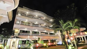 Ein mehrstöckiges Hotel mit Balkonen, einem gut beleuchteten Eingangsbereich und einem angelegten Garten mit Palmen.