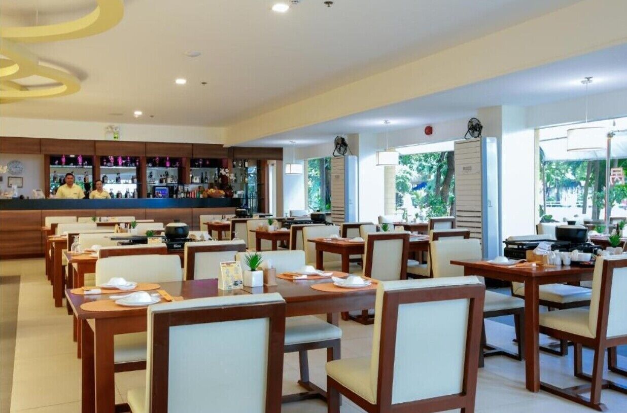 A restaurant with wooden tables and chairs, a bar area, and large windows with a view of greenery.