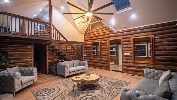 A log cabin interior with a staircase, two grey sofas, a round coffee table, and a ceiling fan. - Timber Lodge: Hot Tub, Fireplace & Fun for All ! (West Milford)