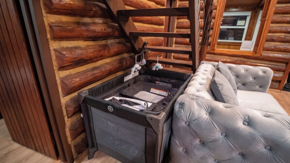 A log cabin interior with a tufted grey sofa, a small black suitcase, and a wooden chair. - Timber Lodge: Hot Tub, Fireplace & Fun for All ! (West Milford)