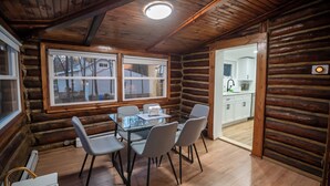 A log cabin interior with a dining area, a kitchen visible through an open door, and a large window. - Timber Lodge: Hot Tub, Fireplace & Fun for All ! (West Milford)
