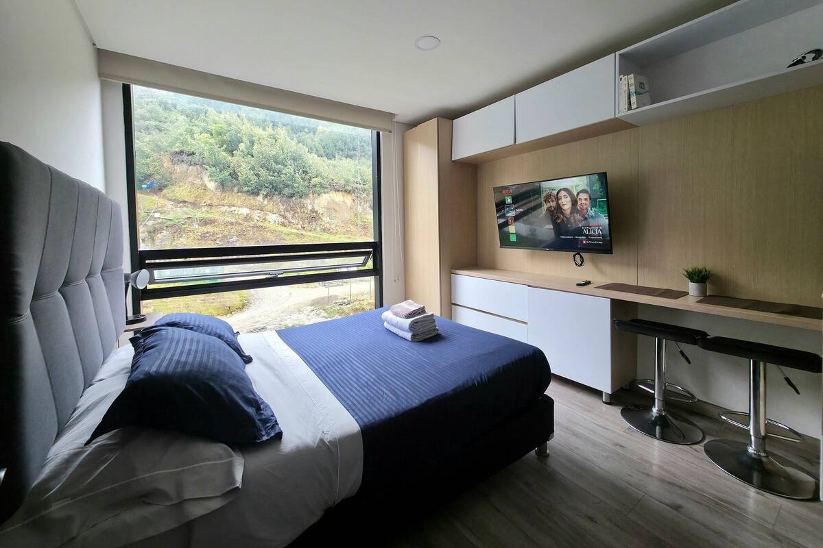 A modern bedroom with a large bed, a TV mounted on the wall, and a view of the outdoors.