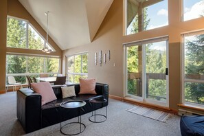 Un moderno salón con un sofá de cuero negro, dos mesas redondas de centro y una zona de comedor con mesa y sillas. Amplias ventanas ofrecen vistas a árboles y un cielo despejado.