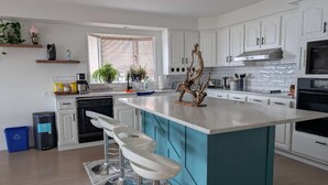 Une cuisine moderne avec des meubles blancs, un îlot central doté de tabourets, un four encastré et une fenêtre avec des stores.