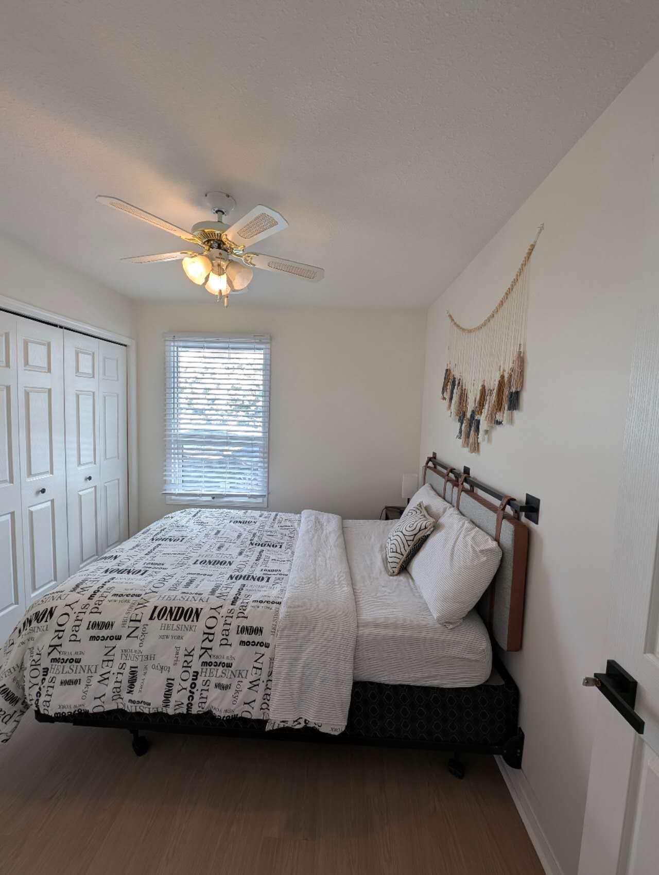 Une chambre à coucher comprenant un lit, un ventilateur de plafond, une fenêtre avec des stores, une décoration murale en macramé et une porte.