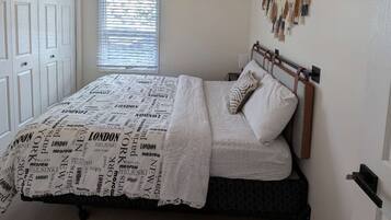Une chambre à coucher comprenant un lit, un ventilateur de plafond, une fenêtre avec des stores, une décoration murale en macramé et une porte.