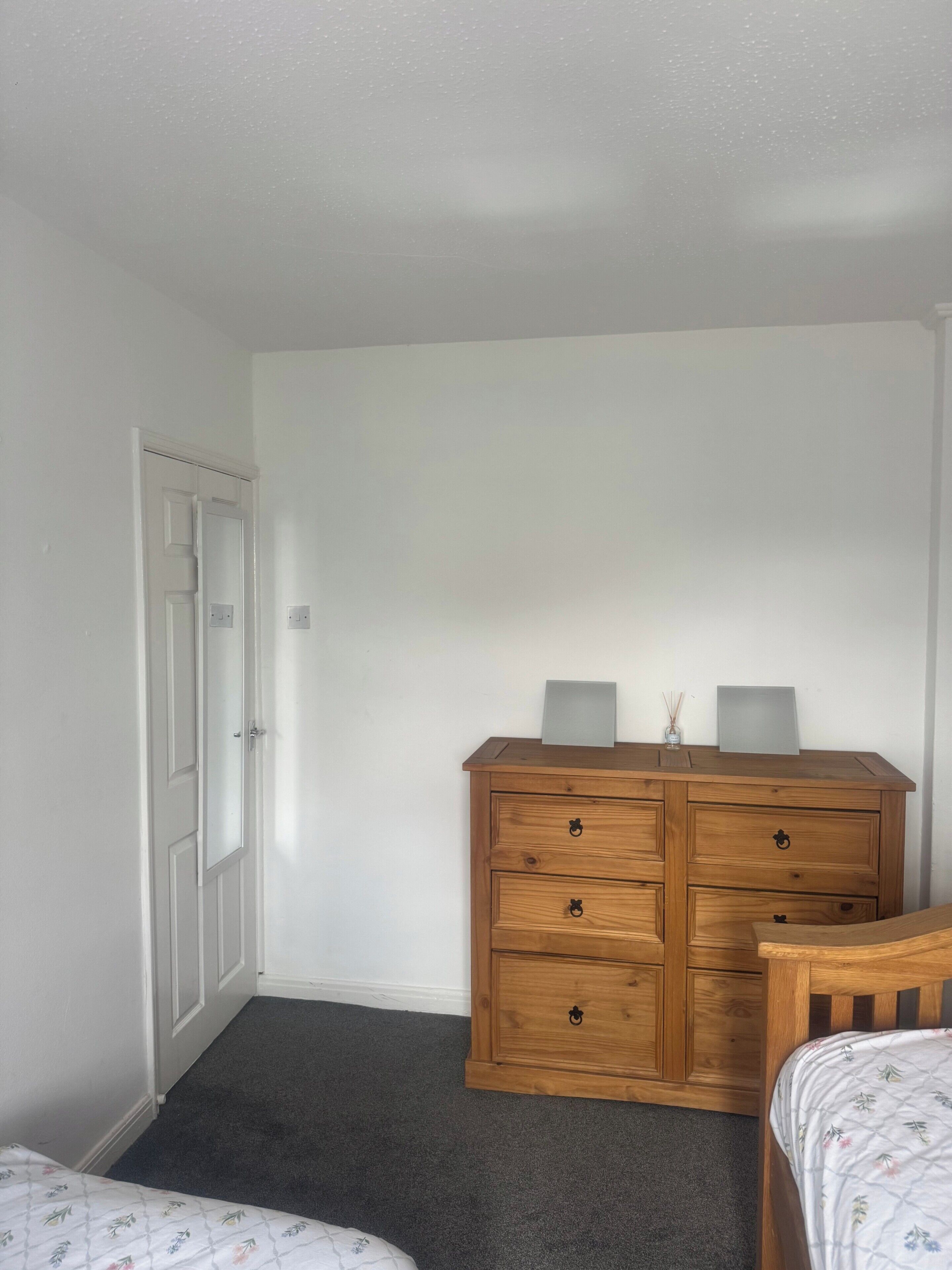 Une chambre avec une commode en bois, un lit recouvert d’une couvre-lit à motifs floraux et une porte blanche.