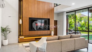 Ein modernes Wohnzimmer mit einem großen Flachbildfernseher an einer Holzwand, einem beigefarbenen Sofa, einem kleinen weißen Tisch und einer Topfpflanze.