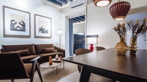 Ein modernes Wohnzimmer mit einem braunen Sofa, einem runden Couchtisch und einem Esstisch mit Stühlen. An der Wand hängen zwei gerahmte Kunstwerke, eine Stehlampe und eine dekorative Vase mit getrockneten Blumen.