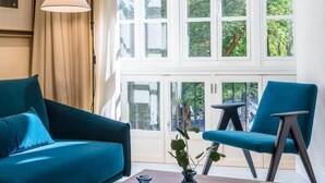 Una habitación con un sofá azul verdoso, un sillón a juego, una mesita pequeña y una lámpara de pie. Amplias ventanas con múltiples vidrios ofrecen vistas a zonas verdes exteriores.