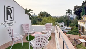 Una terraza en la azotea con sillas blancas y una mesa pequeña, un letrero que dice Refinadores Apartments, y vistas de edificios y árboles circundantes.