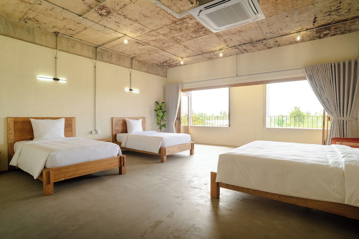 Habitación de hotel con tres camas de madera, un ventanal con vistas y un techo con vigas a la vista y un equipo de aire acondicionado.