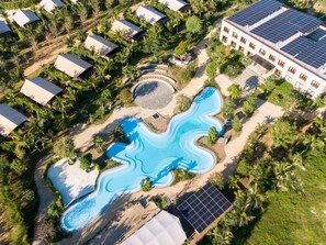 Vue aérienne d’un complexe hôtelier doté d’une piscine à la forme originale, entourée de palmiers et de panneaux solaires.