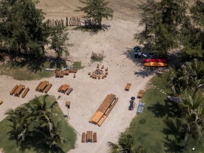 Vue aérienne d’un complexe hôtelier situé en bord de mer, avec des chaises longues en bois, un foyer central et des palmiers.
