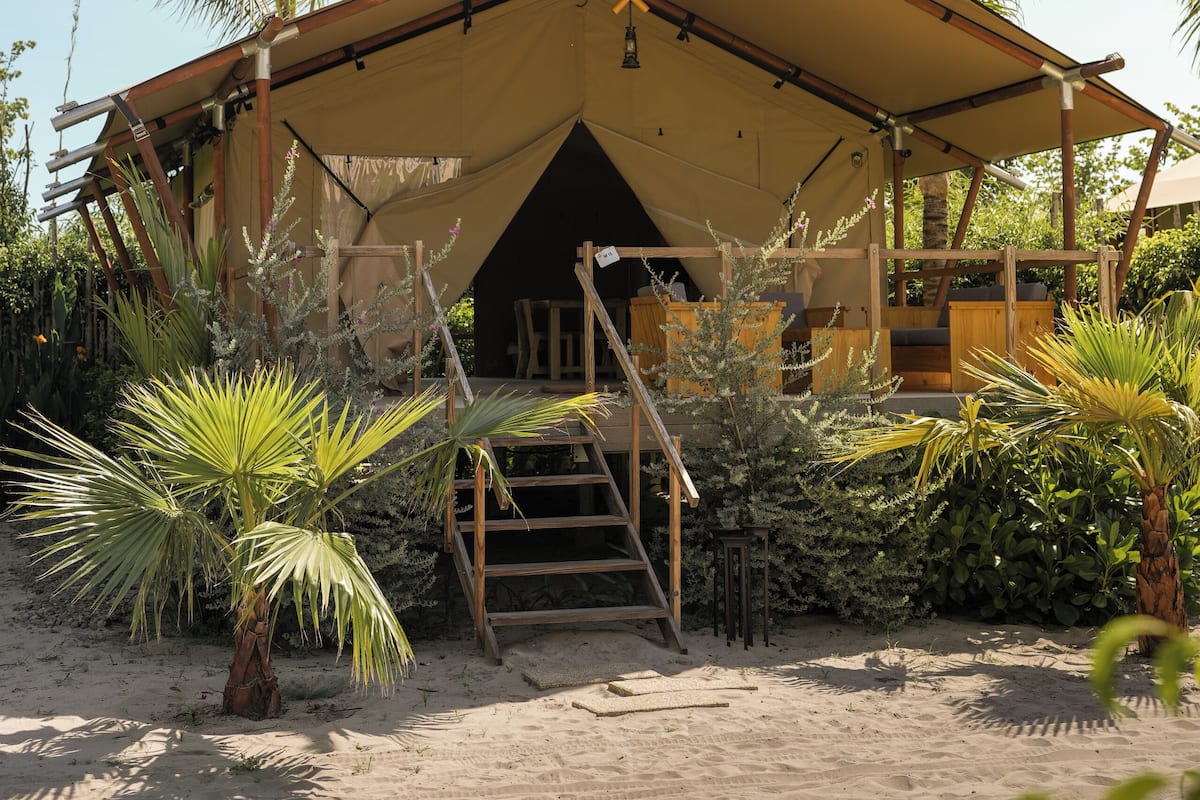 Alojamiento tipo tienda de campaña frente a la playa, con suelo de arena, palmeras y una escalera de madera que lleva a la entrada.