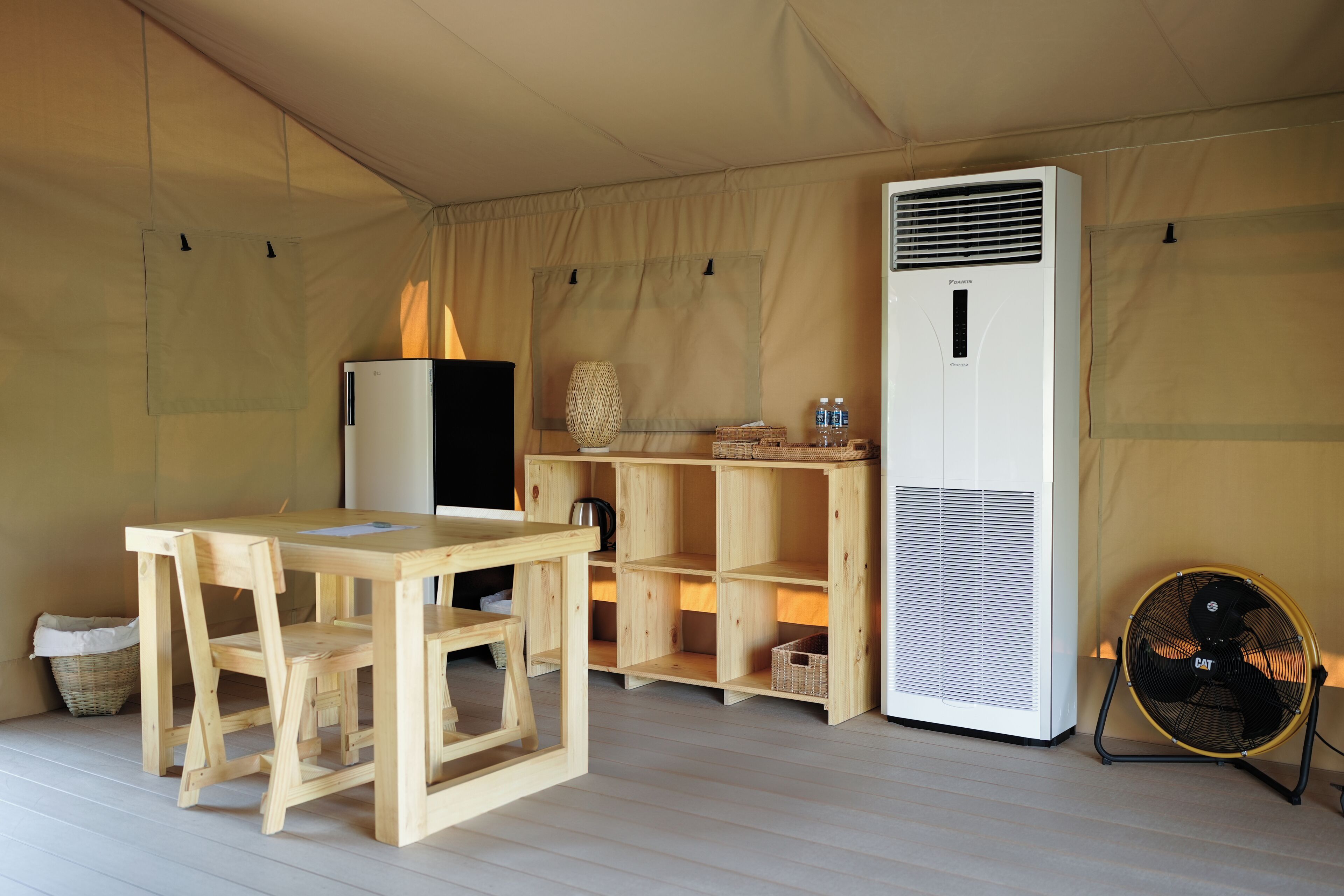 Una habitación con carpa, una mesa de madera y sillas, un refrigerador, un aire acondicionado blanco y un ventilador.