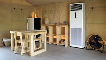 Una habitación con carpa, una mesa de madera y sillas, un refrigerador, un aire acondicionado blanco y un ventilador.