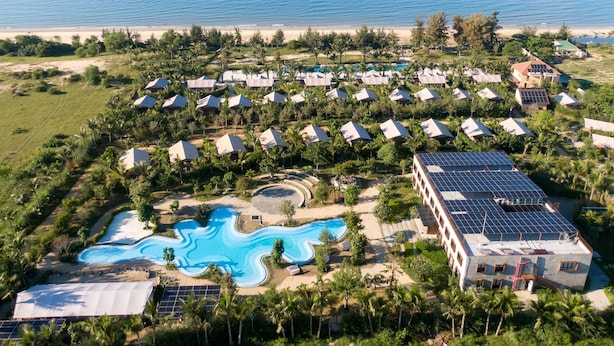 An aerial view of a resort with multiple bungalow-style accommodations, a central swimming pool, and a view of the ocean.
