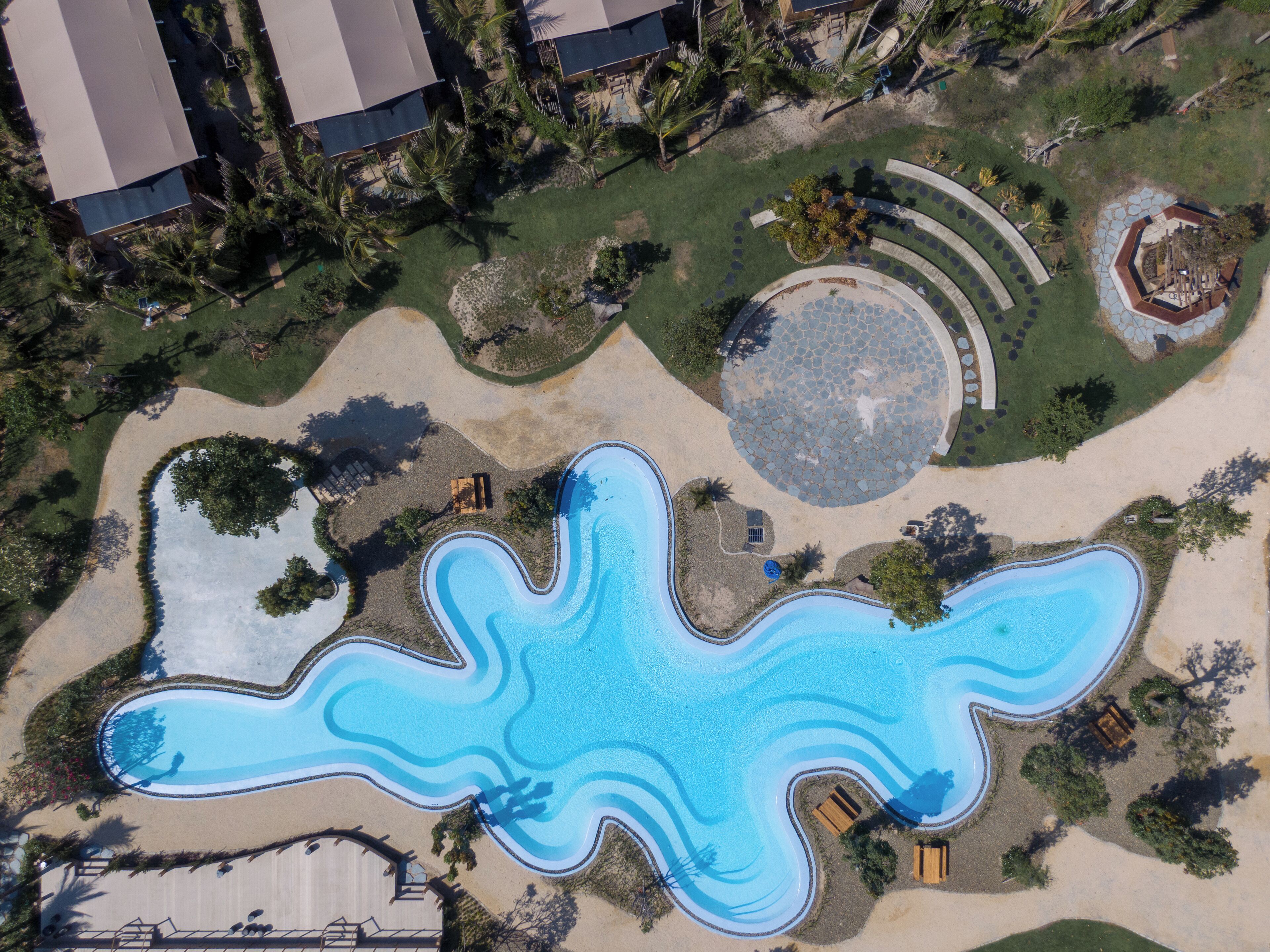 Eine Vogelperspektive zeigt einen luxuriösen Komplex mit einem großen, geschwungenen Pool, umgeben von Palmen und einer runden Steinterrasse.