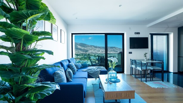 A modern living room with a blue sofa, a coffee table, and a view of a cityscape through large glass doors. - Diamond Ocean View by LovelyStay (Funchal)