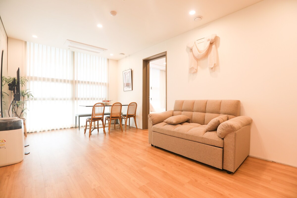 A living room with a beige sofa, wooden dining table, chairs, and a potted plant.