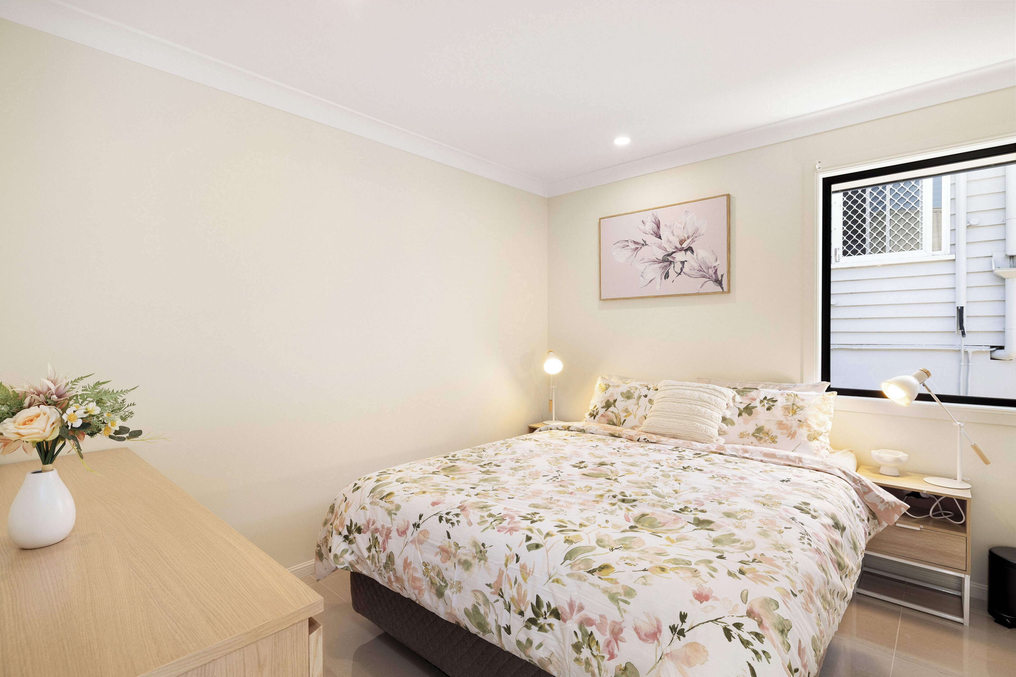 A bedroom with a floral bedspread, a bedside table with a lamp, and a window with blinds.