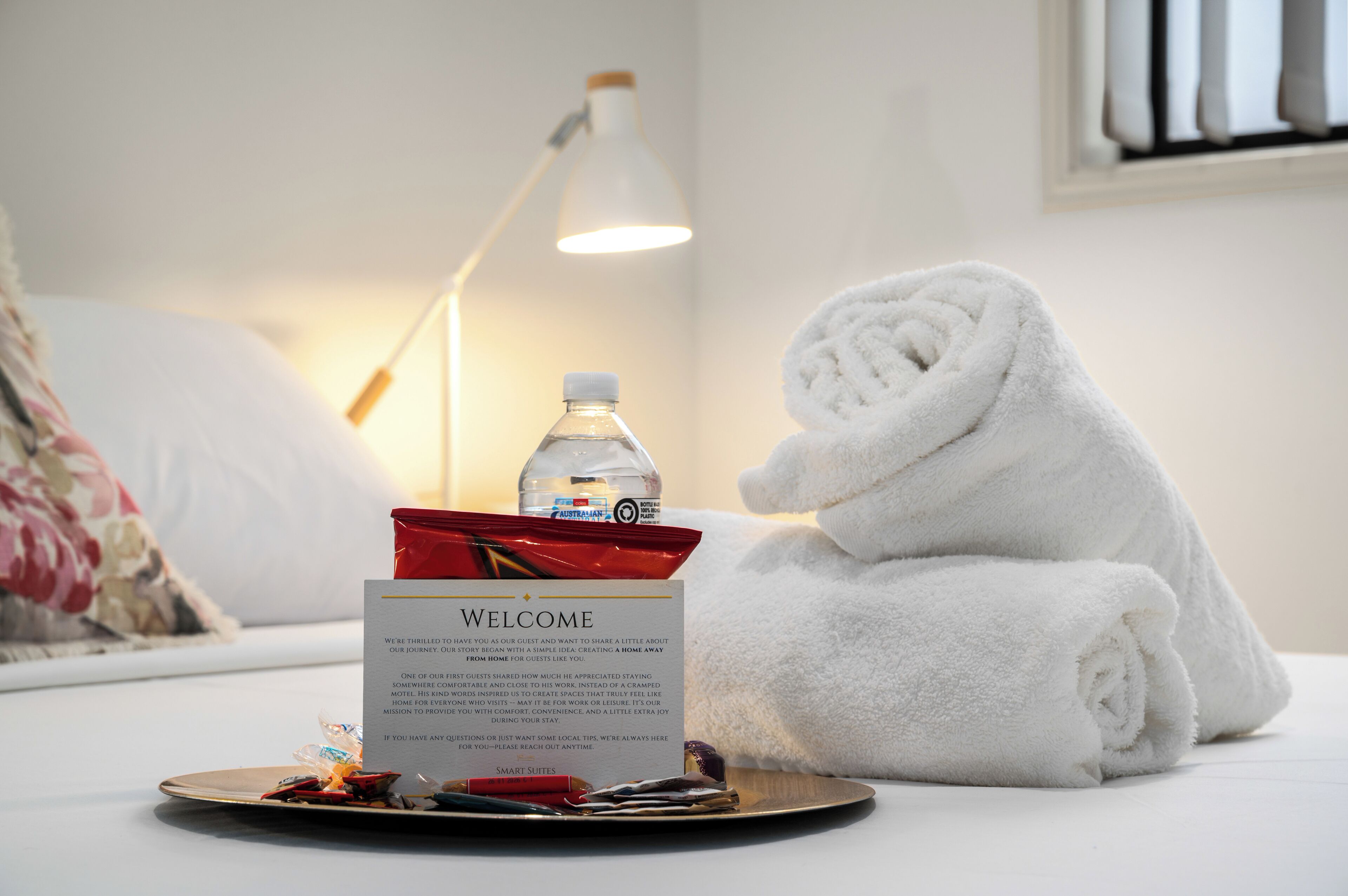 Ein ordentlich bezogenes Bett mit einem gefalteten Handtuch, einer Willkommenskarte, einer Wasserflasche und einem Tablett mit einer roten Tasse und verschiedenen Gegenständen in einem Hotelzimmer.